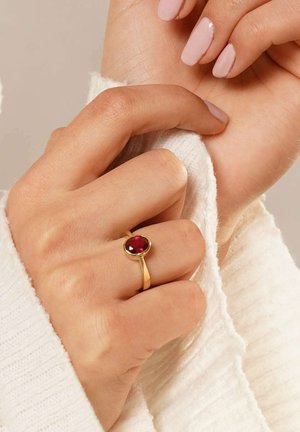 Gold ring with a round red stone, featuring a smooth band. Hand is partially covered by a light-colored, textured fabric.