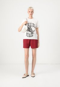 Witte graphic t-shirt met zwarte tekst, rode shorts, beige platte schoenen en een kleine witte tas. Glad textuur en casual ontwerp.