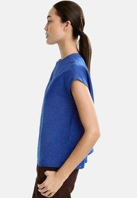 Niebieska lniana bluzka z krótkim rękawem o luźnym fasonie, okrągłym dekolcie i subtelnymi bocznymi rozcięciami, w połączeniu z brązowymi spodniami.