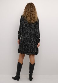 Robe noire avec un motif blanc ondulé, manches longues et ourlet à volants. Associée à des bottes en cuir noires montant jusqu'aux genoux. Tissu doux et fluide.