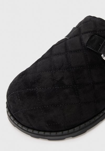 Black quilted suede shoe with rounded toe and rubber sole, featuring a small black strap and silver buckle detail.