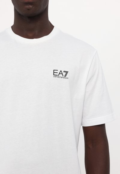 Biała koszulka bawełniana z okrągłym dekoltem i krótkimi rękawami, z czarnym logo "EA7 Emporio Armani" na lewej stronie klatki piersiowej.