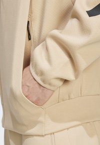 Jaqueta atlética bege com tecido texturizado, bolso lateral com zíper, punhos elásticos e um corte solto. Foco na funcionalidade e conforto no design.