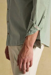 Zeleno-biele pruhované tričko s dlhým rukávom má zapínanie na gombíky, vyhrnuté manžety a decentnú štítok s logom blízko dolného lemu. Hladká textúra látky.