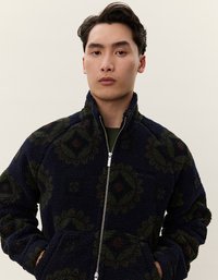 Marineblå fleecejakke med grønne blomstermønstre, med høy krage, glidelås foran og lommer. Myk tekstur, avslappet passform.