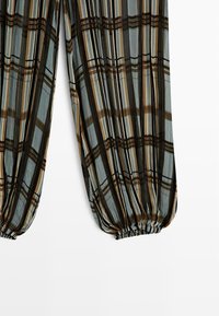 Gennemsigtige bukser med elastiske manchetter, der har et lodret og vandret ternet mønster i sort, brunt og beige på en lyseblå baggrund.