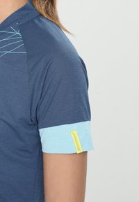 Krátký rukáv košile tmavě modré barvy se světle modrými akcenty. Vyznačuje se strukturální tkaninou a žlutou nášivkou na manžetě. Výrazné detaily šití.