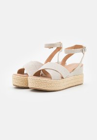 Sandales à plateforme avec des brides en tissu beige et une semelle en jute tissée de style espadrille. Comprend une bride de cheville et un design croisé.
