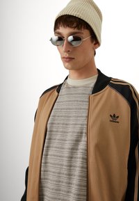 Hiekanvärinen vetoketjutakki mustilla yksityiskohdilla ja Adidas-logo, kerroksittain harmaan raidallisen t-paidan päällä; yhdistetty pyöreisiin hopeisiin aurinkolaseihin ja beigenväriseen pipoon.