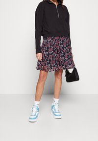 Svart croppad sweatshirt med dragkedja, blommönstrad marinblå kjol i röd och grön, vita ankelsockor, ljusblå sneakers och en liten svart väska.