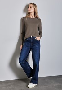 Top a maniche lunghe verde oliva abbinato a jeans a gamba larga blu e sneakers bianche. Tessuto morbido con vestibilità rilassata, dotato di quattro tasche e bottoni.