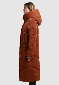 Cappotto lungo imbottito di colore arancio rustico con un ampio cappuccio. Caratterizzato da tasche con cerniera, chiusura con bottoni a pressione e motivo trapuntato per l'isolamento.