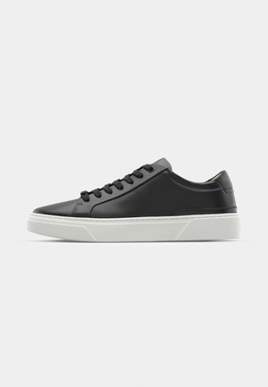 Black leather sneaker with rounded toe, textured surface, white rubber sole, and black laces. Minimalist design with a sleek profile.