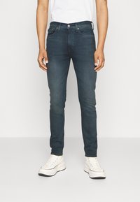 Mörkblå denimjeans med slim fit, fem fickor och knäppning framtill, som bärs med vita sneakers. Slät textur.