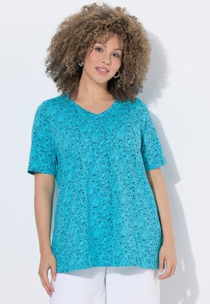 Frau mit lockigem Haar, die ein blau gemustertes Kurzarmoberteil und weiße Hosen trägt, steht vor einem einfachen hellen Hintergrund.