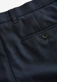 Pantalons marins en tissu lisse, coupe slim, avec une poche arrière fermée par un bouton noir unique, et une texture diagonale subtile.