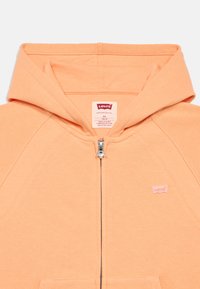 Πορτοκαλί φούτερ με φερμουάρ κατασκευασμένο από μαλακό ύφασμα. Διαθέτει ραβδωτό σχέδιο κοντά στους ώμους και ένα μικρό λογότυπο της Levi's στο στήθος.