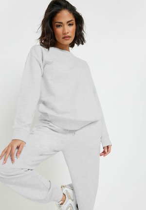 Kobieta z ciemnymi włosami do ramion, ubrana w jasnoszarą bluzę i pasujące spodnie dresowe, pozująca na jednolitym białym tle.