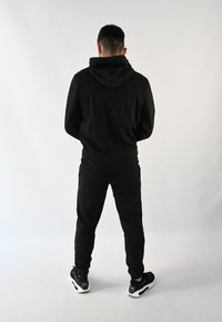 Sudadera negra con capucha y pantalones jogger a juego, con una tela texturizada. Zapatillas negras con acentos blancos completan el atuendo monocromático.