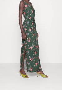 Blommig maxiklänning i grönt tyg med V-ringning, insvängd midja och sidofrackar; prydd med rosa och blå blomster mönster. Matchas med gröna klackar.