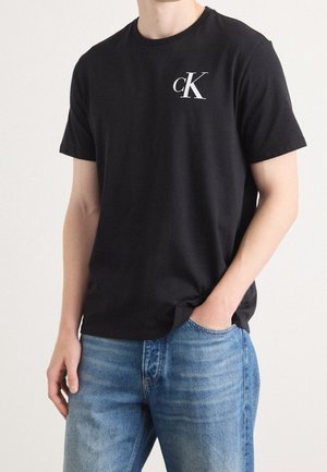 Mężczyzna w czarnej koszulce z krótkim rękawem z białym logo "cK" na piersi i niebieskich dżinsach, jedna ręka w kieszeni.