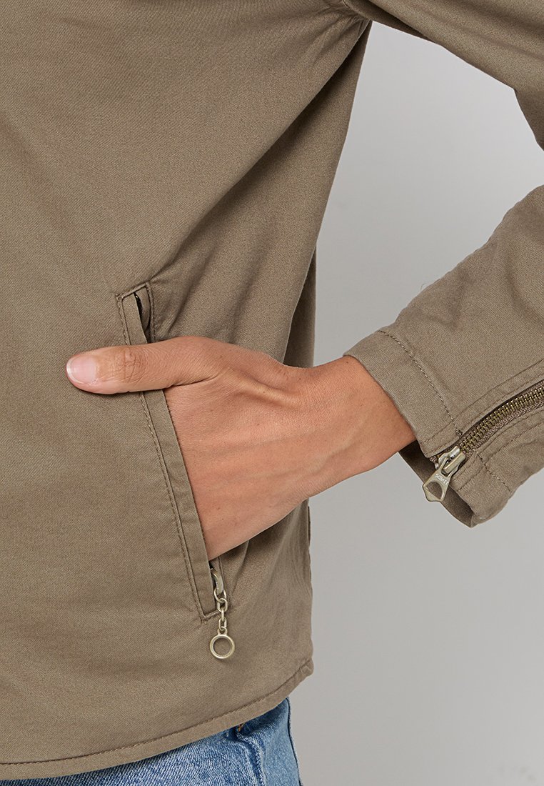 Veste en coton marron avec une poche latérale et une fermeture éclair sur la manche. Le tissu est lisse, et le design présente des détails de couture minimalistes.