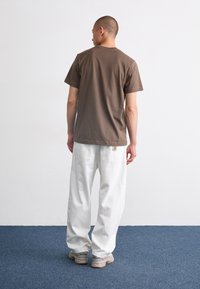 Brun bomull T-skjorte med korte ermer, kombinert med løstsittende hvite bukser og grå sneakers. Enkelt design, ingen synlige mønstre.