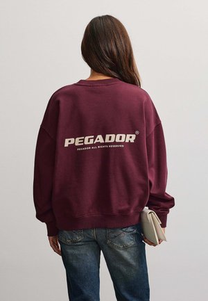 Persona con cabello largo y castaño lleva una sudadera burdeos con el texto "PEGADOR" en la espalda, vaqueros azules y sostiene un bolso beige plegado.