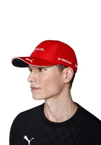 Czerwona czapka z lekko wygiętym daszkiem, z czarnym wzorem w kratkę. Widać loga: Porsche, TAG Heuer i Puma. Noszona z czarną koszulą.