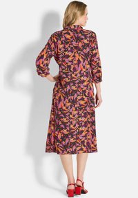 Maxikleid mit floralem Muster in den Farben Pink, Orange und Schwarz, ausgestattet mit langen Ärmeln, einem kragenförmigen Ausschnitt und einer lockeren Passform. Rote Blockabsatzsandalen.