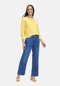 Geel gemêleerd sweatshirt met wijde halslijn, gecombineerd met blauwe wijde jeans en gele haksandalen. Model staat voor een effen achtergrond.