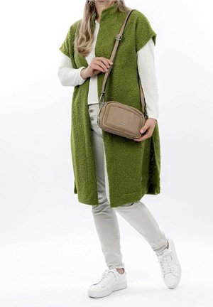 Kobieta w długiej zielonej teksturowanej kamizelce, białej koszuli z długim rękawem, jasnych spodniach, białych trampkach, trzymająca beżową torbę na ramię z plecionym detalem.
