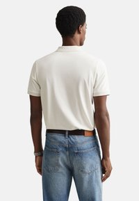 Bela polo majica s kratkimi rokavi in klasičnim ovratnikom, kombinirana s svetlo modrimi jeans hlačami in temno rjavim pasom. Preprost, čist dizajn.