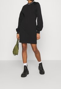 Svart sweatshirtklänning med puffärmar, ribbat halsringning och logotypdetalj. Matchad med grön väska och svarta kraftiga ankelstövlar.