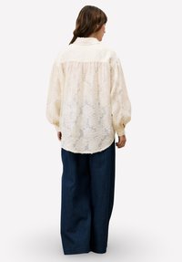 Crèmekleurige, doorschijnende blouse met bloemenkantpatroon; hoge halslijn en losse pasvorm. Gecombineerd met wijde, donkerdenim broek.