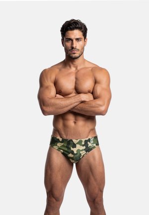 Muskularny mężczyzna ze skrzyżowanymi ramionami, ubrany w zielone kąpielówki w kamuflaż, stojący na jasnym, jednolitym tle.