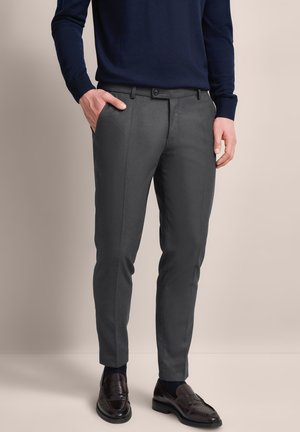 Graue maßgeschneiderte Hosen mit flacher Vorderseite, schlankem Schnitt, aus glattem Stoff. Kombiniert mit einem marineblauen Pullover und dunklen Schuhen.
