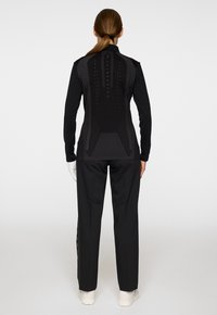 Vrouw met een bruin paardenstaart, gekleed in een zwart longsleeve top met een patroon op de achterkant, zwarte broek, witte handschoenen en witte schoenen tegen een witte achtergrond.