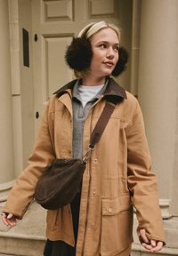 Chaqueta de lona marrón con un cuello oscuro, camiseta polo gris y bolso cruzado marrón. Los auriculares de piel sintética negros añaden detalle al conjunto.