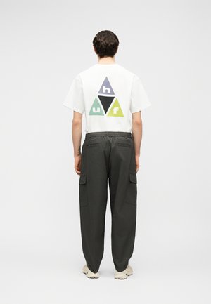 Άτομο που στέκεται πλάτη προς το φακό, φοράει λευκό πουκάμισο με πολύχρωμο τρίγωνο λογότυπο και σκούρο παντελόνι cargo με αθλητικά παπούτσια.