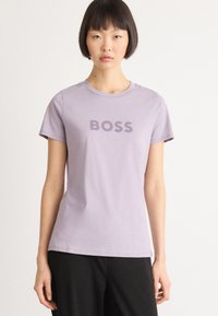 Jauna moteris su trumpais tamsiais plaukais, apsirengusi alyvų spalvos marškinėliais su užrašu "BOSS" priekyje ir juodomis kelnėmis, stovi prieš paprastą foną.