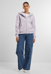 Felpa zip-up lavanda con tasca a marsupio, abbinata a pantaloni cargo in denim blu larghi con tasche laterali e fondo dritto.