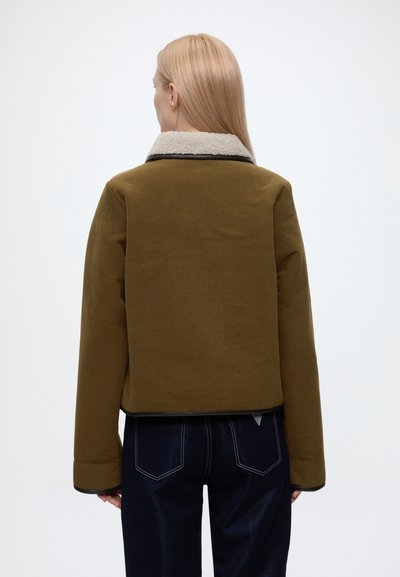 Femme aux cheveux blonds raides porte une veste vert olive avec un col en polaire blanc et un jean bleu foncé, vue de dos.