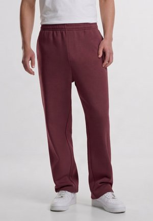 Homme portant un pantalon de survêtement bordeaux à taille élastique et des baskets blanches, debout devant un fond clair uni.