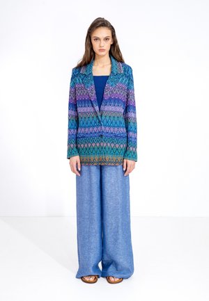 Geweven blazer in een multi-kleuren geometrisch patroon met tinten blauw, paars en groen; gecombineerd met wijde lichtblauwe broeken.