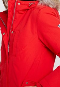 Rode winterjas met een ritssluiting aan de voorkant, een bont gevoerde capuchon en zijzakken. Heeft een gladde stof en een verstelbaar trekkoord bij de kraag.