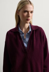 Bordeaux gebreide trui met een V-hals en brede kraag, gelaagd over een blauw-wit geruit shirt, toont soepel materiaal en een relaxte pasvorm.