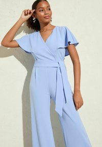 Combi-short bleu clair avec des manches volantées et une taille ceinturée. Tissu lisse, jambes larges et décolleté en V plongeant.