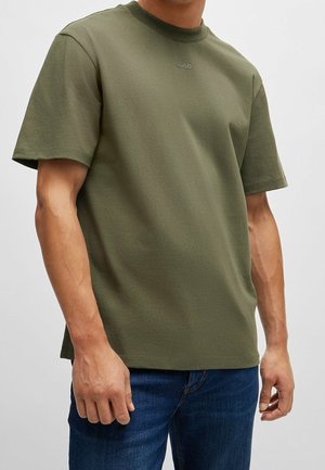 Mand iført olivengrøn kortærmet t-shirt med lille "HUGO" logo på brystet og blå jeans mod en ensfarvet baggrund.