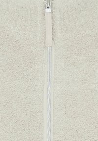 Beige Fleecejacke mit Reißverschluss, hergestellt aus weichem, strukturiertem Stoff, mit sichtbarem weißem Reißverschluss und passendem Zipper-Anhänger.